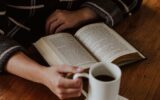 Person leser en åpen bok ved et trebord med en kopp kaffe