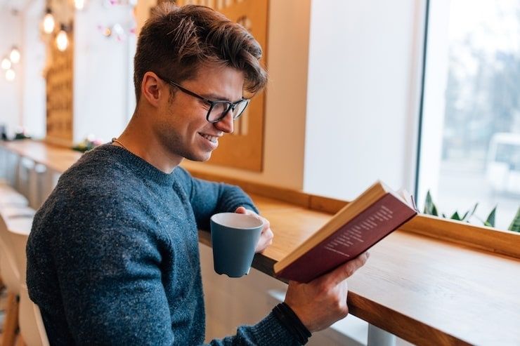 Smilende mann leser en bok med en kopp kaffe ved vinduet